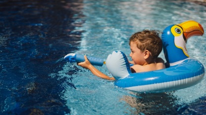 A boy with a joyful expression is floating in a swimming pool on a sunny day, using an swimming circle designed to look like a blue bird.