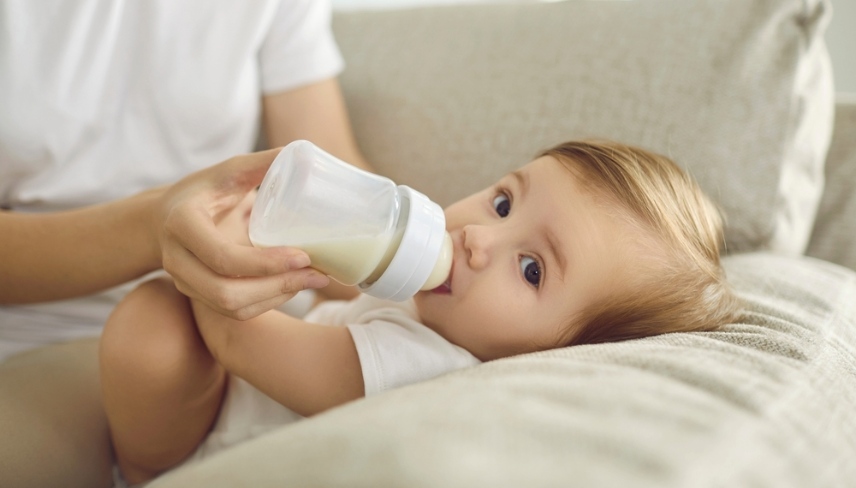 Baby enjoying nutritious meal. Mother feeding baby on sofa. Nanny formula feeding little child as alternative to breastfeeding. Baby boy or girl lying on couch and drinking milk from plastic bottle