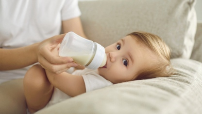 Baby enjoying nutritious meal. Mother feeding baby on sofa. Nanny formula feeding little child as alternative to breastfeeding. Baby boy or girl lying on couch and drinking milk from plastic bottle