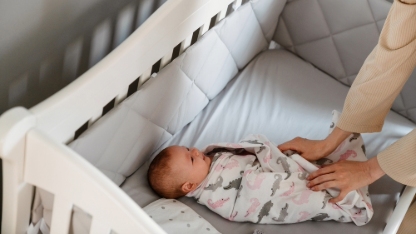 White young woman in housecoat swaddling her newborn baby in crib at bedroom