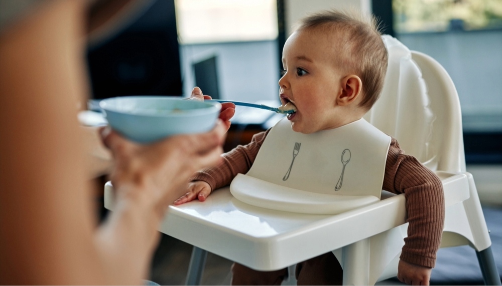 Unrecognized woman feeding her baby son sitting in a high chair.