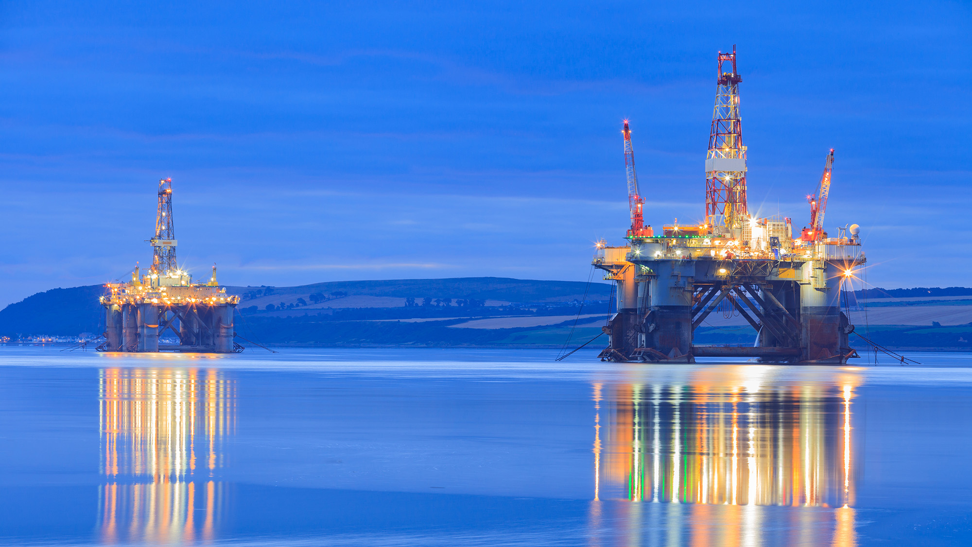 Two illuminated offshore oil rigs stand in calm water at dusk, with bright industrial lights reflecting across the smooth blue surface under a cloudy evening sky.